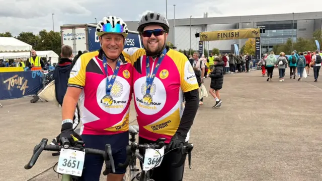 Dos hombres con ropa de ciclismo sonríen a la cámara en un evento benéfico. Sus bicicletas llevan el dorsal en la parte delantera y se abrazan.