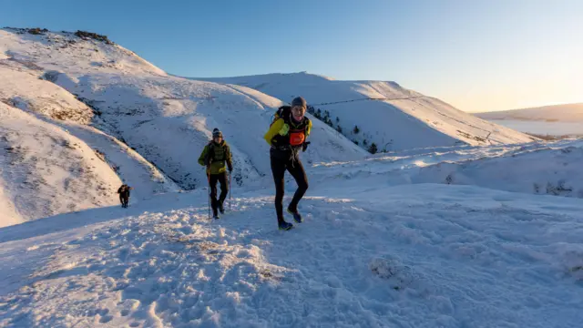 Tres corredores ascienden por laderas nevadas en pleno invierno. Llevan ropa de abrigo. Es un día soleado y parece que va a amanecer.
