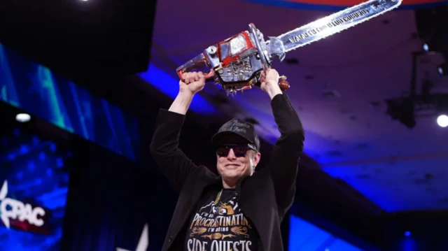 Elon Musk Muhafazakar Siyasi Eylem Konferansı'nda sahnede, bürokrasideki kesintileri sembolize etmek için kullandığı bir testereyi havaya kaldırıyor. Siyah gözlük ve siyah şapka takıyor.