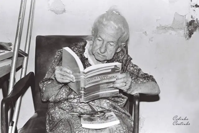Cora Coralina sentada numa cadeira lendo