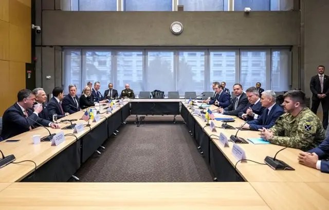 Dikdörtgen şekilde birleştirilmiş dört masa etrafında takım elbiseli erkek ve kadınlar ve bir de kamuflajlı general oturuyor. 23 Kasım 2025 Cenevre. 