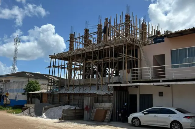 Uma obra em andamento ocupa a frente de um imóvel, com uma estrutura de concreto e madeira sendo construída. Ao lado, há uma casa já pronta, um carro estacionado e materiais de construção espalhados pela calçada, em uma rua de área urbana.