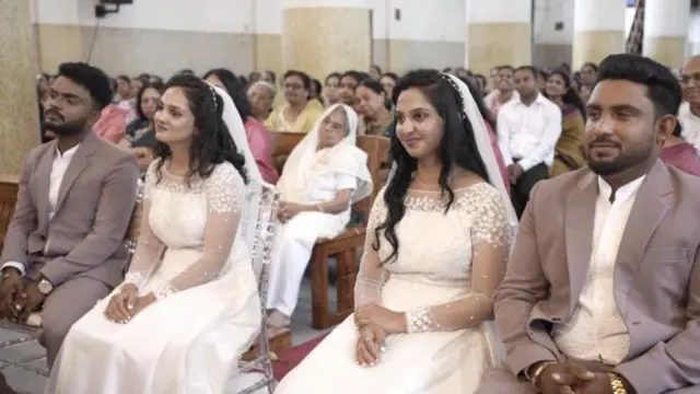 Nikhil Thomas-Maria Mary (left) and Magi Mary-Akhil Thomas (right) in their wedding gowns sitting side by side inside a church, with guests in the background