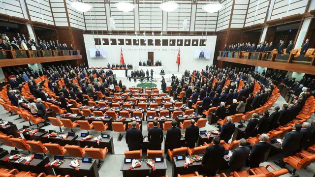 TBMM Genel Kurulu'ndaki milletvekilleri ayakta bekliyor. Arkalarında turuncu sandalyeler ve kahverengi masalar görünüyor. Kürsünün arkasında beyaz arka plan üzerine 'Egemenlik Kayıtsız Şartsız Milletindir" yazıyor.