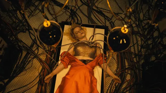 Дівчина в помаранчевій сукні лежить на ліжку, до неї підключені дроти