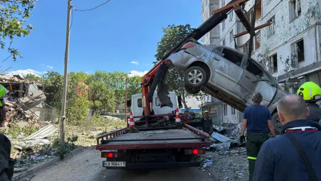 Троє рятувальників спостерігають, як пошкоджений автомобіль підіймають на платформу вантажівки.