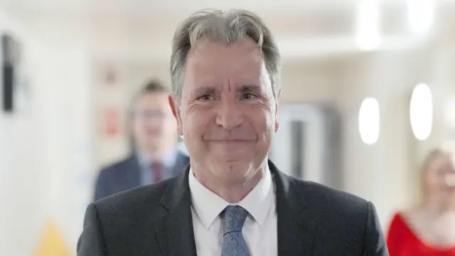 Fotografía del diputado Dan Norris con traje y corbata. Al fondo se ven dos personas que no aparecen en foco.