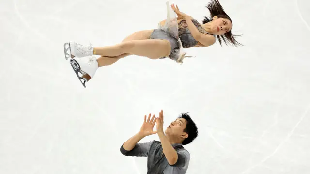 Японские фигуристы Рику Миура и Рюити Кихара: девушка в полете, юноша держит руки поднятыми после того, как он ее подкинул вверх
