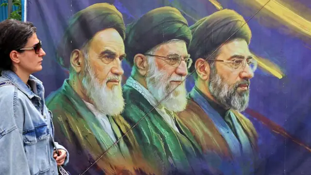 تصویری از یک زن در کنار نقاشی روح الله خمینی، علی خامنه‌ای و مجبتی خامنه‌ای