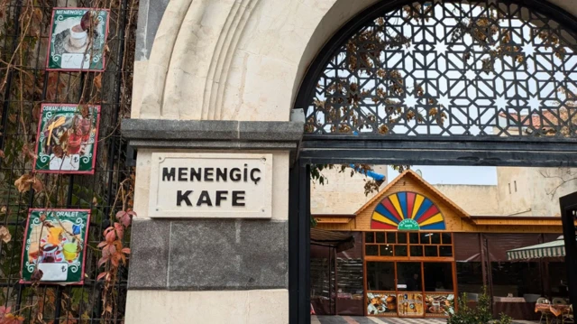 Wejście do Menengiç Kafe, tradycyjnej kawiarni w Gaziantep. Na pierwszym planie kamienny filar - na nim kamienna tablica z nazwą kawiarni - i metalowe zdobienia nad wejściem. Na lewo od wejścia kilka kolorowych plakatów reklamujących podawane w kawiarni napoje. Na dalszym planie widać dziedziniec, na nim stoły dla klientów i drewniany budynek z zamkniętymi przeszklonymi drzwiami. W głębi ściany wyższego, kamiennego budynku.