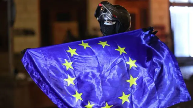 Manifestante con casco de moto, cara cubierta y una bandera de la Unión Europea. 