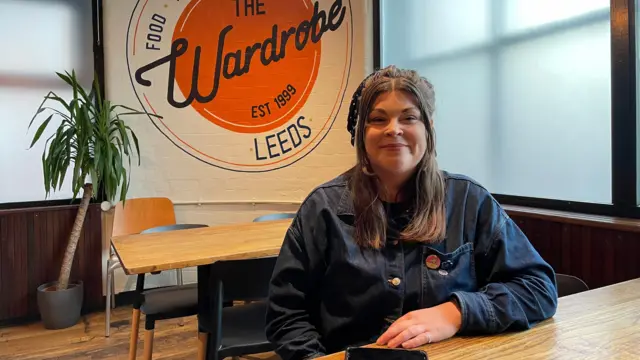 Una mujer con una camisa vaquera azul está sentada a una mesa. Tiene el pelo castaño y sonríe. Detrás de ella, una pancarta dice "El Armario Leeds".