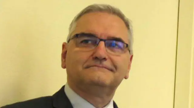 Mężczyzna w ciemnej marynarce, jasnej koszuli i niebieskim krawacie, o krótkich siwych włosach. Ma na sobie okulary. Za nim znajduje się gładka ściana w kolorze kremowym.