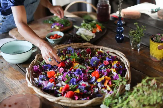 Mesón con una canasta de flores comestibles en primer plano, y otros ingredientes alrededor, con la mano de una mujer que está preparando comida tomando algunas