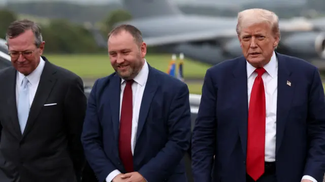 Троє чоловіків зліва направо – Воррен Стівенс, посол США у Великій Британії, міністр охорони здоров'я Шотландії Ієн Мюррей та президент США Дональд Трамп. Вони в аеропорту на розмитому фоні, ймовірно, зображений літак Air Force One. Усі троє в костюмах.