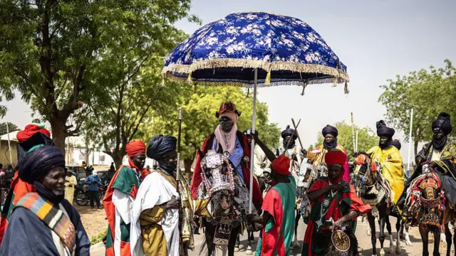 The Emir of Dutse sits on a white horse, shielded by a blue parasol. He is joined by horse riders wearing colorful robes.