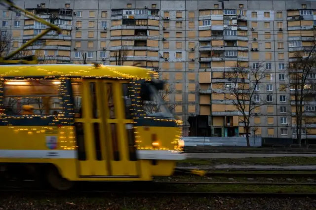 Жовтий трамвай, прикрашений святковими гірляндами з маленьких вогників, що рухається по трамвайних коліях. На задньому плані – багатоповерховий житловий будинок із численними вікнами, більшість з яких забиті дерев’яними дошками. Довкола – голі дерева, а земля виглядає вологою, без снігу.