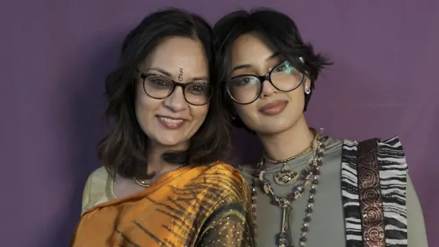 Dos mujeres, una al lado de la otra, sonrientes, con atuendos coloridos. Ambas llevan gafas. La mujer de la izquierda es un poco mayor y la de al lado es más joven, con el pelo más corto. Están frente a un telón de fondo morado.