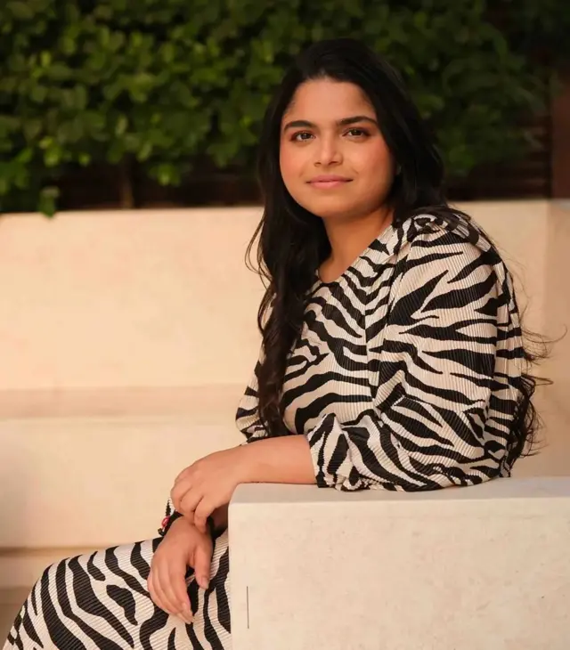 ایتی بهاسین با پیراهنی با خطوط سیاه و سفید روی مبل نشسته و به دوربین نگاه می‌کند