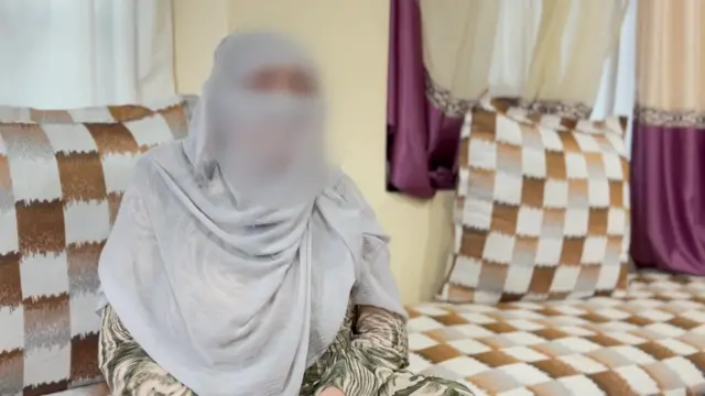 薄いグレーのスカーフを頭部に巻いた女性が、クッションに座っている。女性の顔には、身元を隠すためにモザイクがかけられている