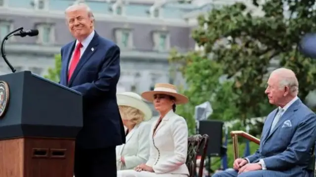 Президент Трамп отводит взгляд от микрофона и улыбается, произнося приветственную речь в честь короля Карла III. Король, первая леди и королева сидят позади него.
