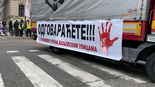 Na belom transparentu prikačenom za kamion ispisano je 'Odgovaraćete za prolivenu krv na valjevskim ulicama sa crvenom šakom'