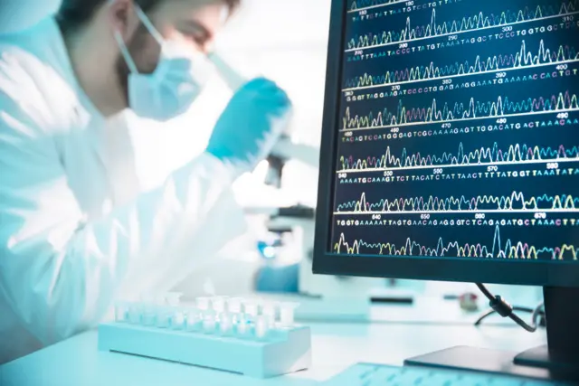 Sequência de DNA na tela de um computador, com pesquisador ao fundo, em um laboratório