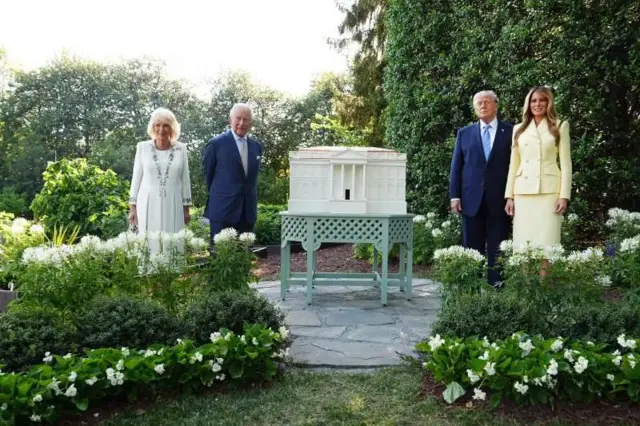 King Charles III and Queen Camilla join US President Donald Trump and First Lady Melania, all dressed formally, in a green garden, beside a beehive in the shape of the White House