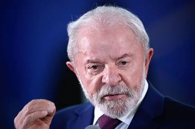 Beyaz sakallı bir adam. Eliyle işaret ederek konuşuyor. Arkasında lacivert bir fon.