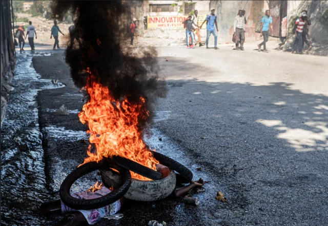 En primer plano, un cadáver arde bajo unas llantas en llamas en una calle del centro de Puerto Príncipe, Haití, mientras al fondo se ven unas personas desenfocadas, el 9 de febrero de 2026. 