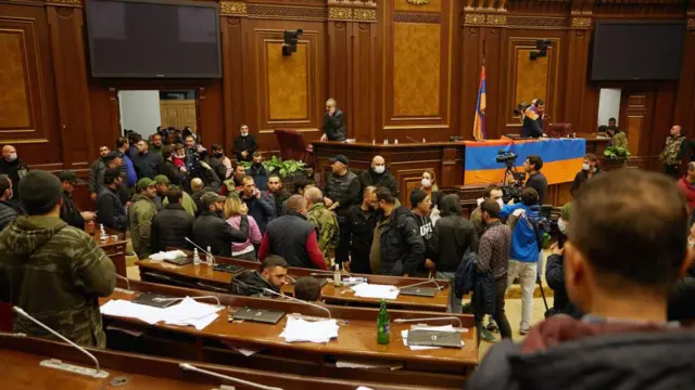 Protestocular ve kamulafjlı kişiler Ermenistan parlamentosu genek kurulunda. Kürsüye bir Ermenistan bayrağı serilmiş durumda. Bazı göstericilerin üzerinde kamuflaj var.
