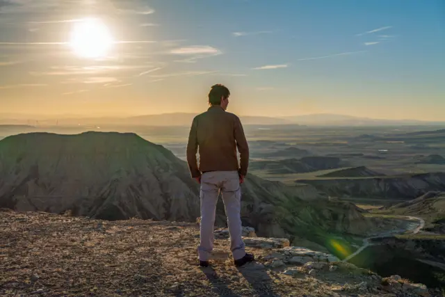 O Prof. Brian Cox olha para o horizonte do topo de uma montanha. Ele está vestindo uma jaqueta marrom e calças cinza. Colinas verdes podem ser vistas à distância, e o Sol brilha intensamente