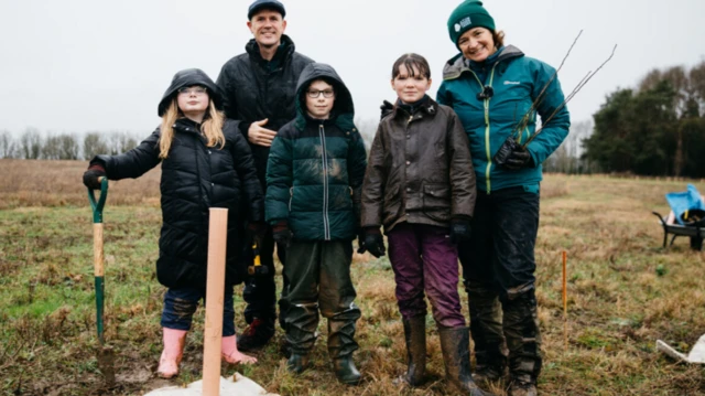 Luke Jerram standing in a field with a woman and three children, one boy and two girls, all in wet weather gear. One of the children is holding a spade upright and there is a sapling covered in plastic in the foreground and a wheelbarrow behind the group