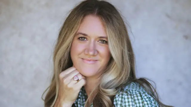 Женщина с длинными светлыми/каштановыми волосами улыбается в камеру. Ее правая рука подпирает подбородок. На безымянном пальце у нее кольцо в форме розового сердца. Она одета в зелено-белую клетчатую рубашку. 