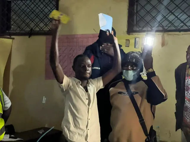 Un homme en chemise claire lève haut les bras, tenant un bulletin jaune dans une main et une enveloppe dans l'autre main. À sa gauche, une femme portant un bonnet brillant, un masque chirurgical et une veste beige et noire, lève également un bras en tenant une lampe allumée qui éclaire la scène.