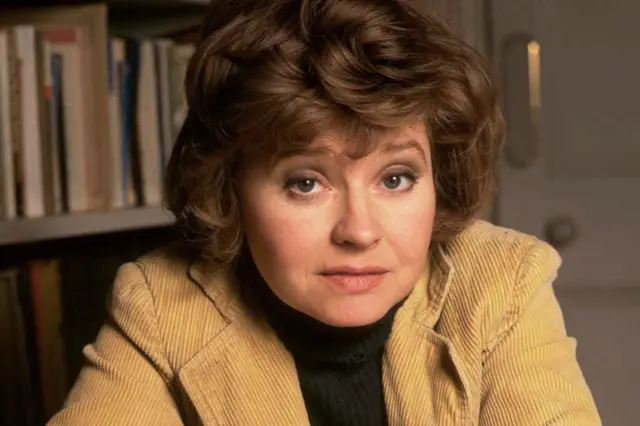 Prunella Scales is wearing a light brown corduroy jacket over a dark turtleneck sweater is seated indoors. Behind her is a bookshelf filled with books, and to the right, part of a light-coloured door with a round handle is visible. The setting appears to be a cozy, study-like environment with warm lighting.
