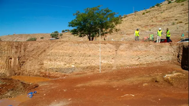 Gran terreno excavado en mitad de la montaña. Se ven varias personas a lo lejos, con chalecos amarillos.