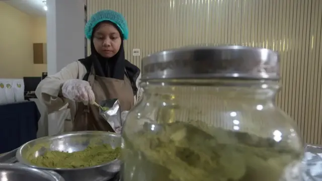 Staf Arafa Tea, yang mengenakan kerudung, celemek, dan sarung tangan, menyendokkan bubuk matcha dari baskom stainless ke dalam kemasan. 