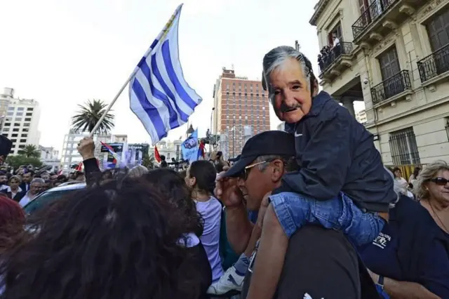 Multidão para receber Mujica, com a bandeira do Uruguai, e uma com uma máscara do ex-presidente uruguaio nas costas de um adulto