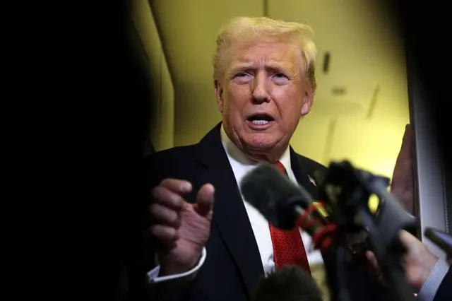 El presidente de Estados Unidos, Donald Trump, habla con la a bordo del Air Force One. En la imagen se le ve con traje de chaqueta azul marino oscuro, camisa blanca y corbata roja, se le solo una de las manos y al lado derecho, varios micrófonos de la prensa.
