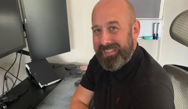 Vêtu d'un t-shirt noir, Andrew Tillman est assis à son bureau et sourit, face à la caméra.