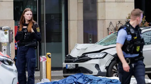Dwóch nieostrych policjantów stoi przed białym samochodem z wybitą szybą i zgiętą maską, przykrytymi niebiesko-białymi plandekami na ziemi.