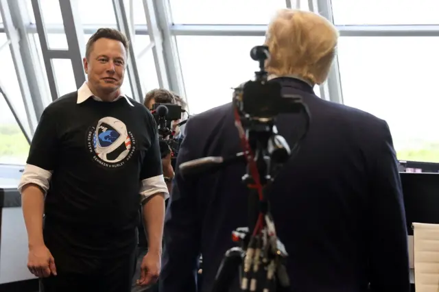 A foto mostra Elon Musk, um homem branco, sem barba, com o cabelo curto, vestindo uma blusa preta com a estampa de um astronauta sobre uma camisa branca. Ele está de frente, enquanto Trump, vestindo um terno azul escuro, está de costas.