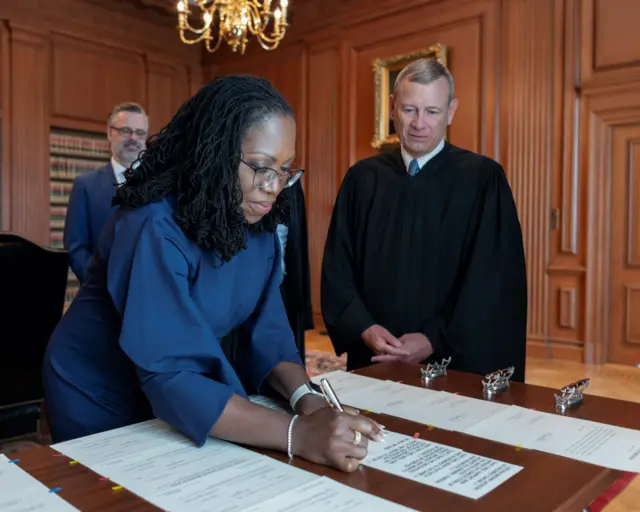 Ministra Ketaji Brown Jackson é uma mulher negra. Ela está assinando um papel. Ao lado dela há um homem em pé