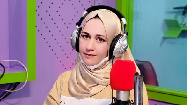 شمین نجرابی در استدیوی رادیو با دیوار سبز و بنفش پشت میکروفون نشسته است. او چادر زرد رنگ به سردارد.