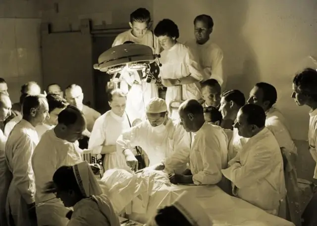 Diveras pessoas de branco em torno em uma mesa de cirurgia onde não é possível ver o paciente, apenas uma figura coberta