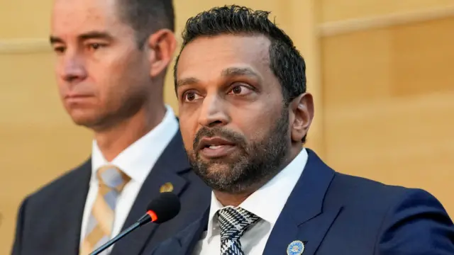 Le directeur du FBI, Kash Patel, s'exprime lors de la conférence de presse. Patel a une barbe claire et porte une veste bleue, une chemise blanche et une cravate à motifs gris, noirs et blancs.
