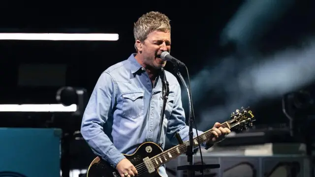 Noel Gallagher em uma camisa jeans azul clara segurando seu violão enquanto canta