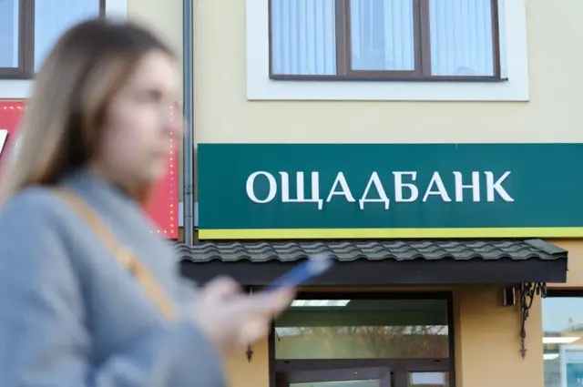 Перед фасадом відділення Ощадбанку з зеленим вивіскою жінка тримає у руках смартфон.