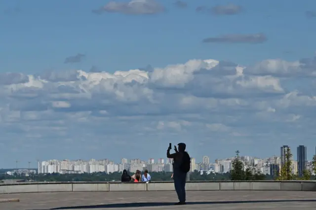 Вигляд на багатоповерхівки лівого берега Києва; на передньому плані чоловік фотографує на телефон.